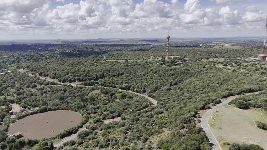 Drone flies over reservoir toward large radio tower on top of Naval Hill on partially cloudy afternoon in Bloemfontein, South Africa