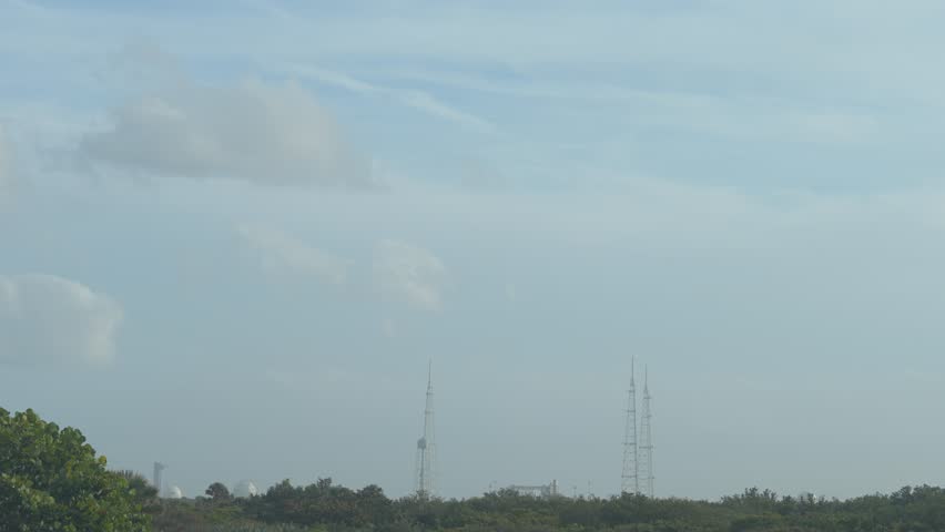 A rocket launches from a coastal space center with communication towers and infrastructure visible under a cloudy sky. Bright engine flames rise above trees and buildings as the spacecraft lifts off