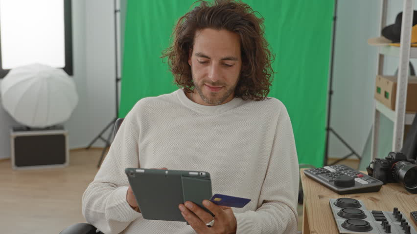 Young hispanic man holding tablet and creditcard with hands visible in studio beside green screen and camera gear; creative contentment.