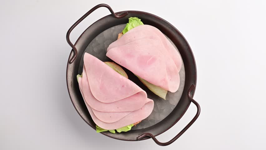 Two sandwiches with layers of ham and lettuce are arranged in a bowl. The sandwiches are placed on a white surface. The setting is simple and focuses on the food.