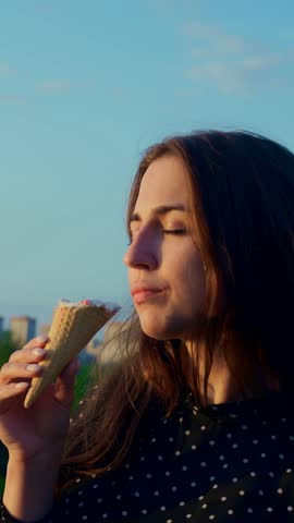 Sunset woman eating ice cream rooftop city skyline background, caucasian student wearing polka dot blouse holds waffle cone, golden hour light warms brunette profile, contemplative expression, slow