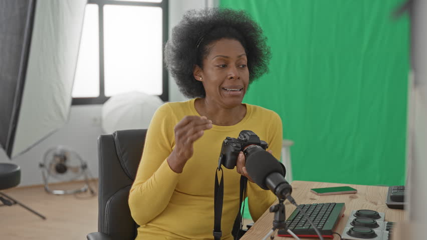 Woman adjusting camera with hand at desk in studio beside microphone and green screen, wearing yellow sweater; concentration creativity.