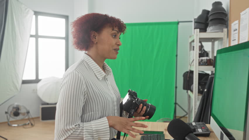Woman holds camera and gestures with hands in studio with green screen and lighting kit; focused teaching.