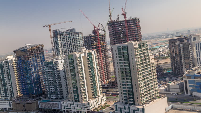 Tall skyscrapers under construction with heavy cranes near the water canal in Dubai Business Bay. Daytime aerial view of fast urban real estate development in time-lapse.