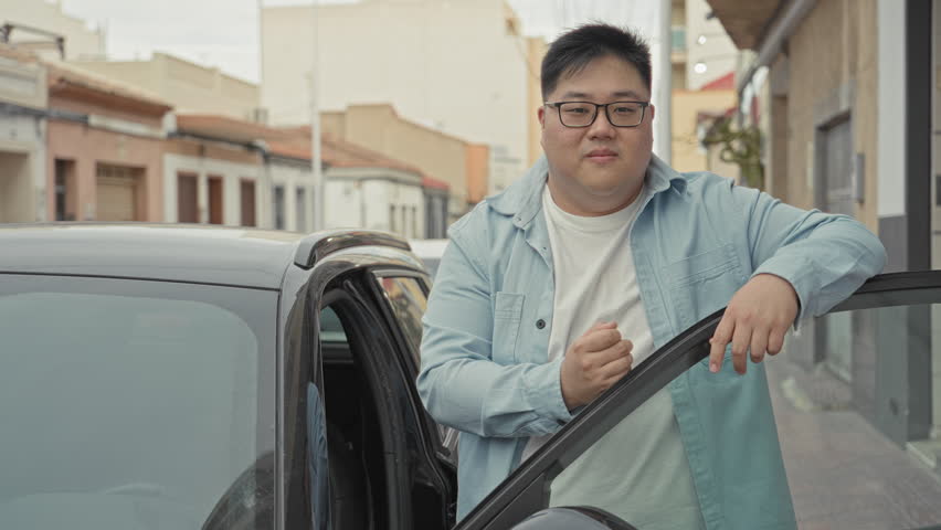 Man holding car key and leaning on open car door while smiling and gesturing to hand keys on a residential street; pride ownership.