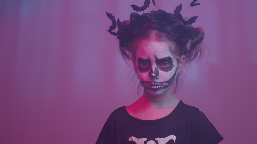 Portrait of little girl wearing skeleton face paint and bat headwear. Child looking at camera with spooky expression creating dark Halloween mood