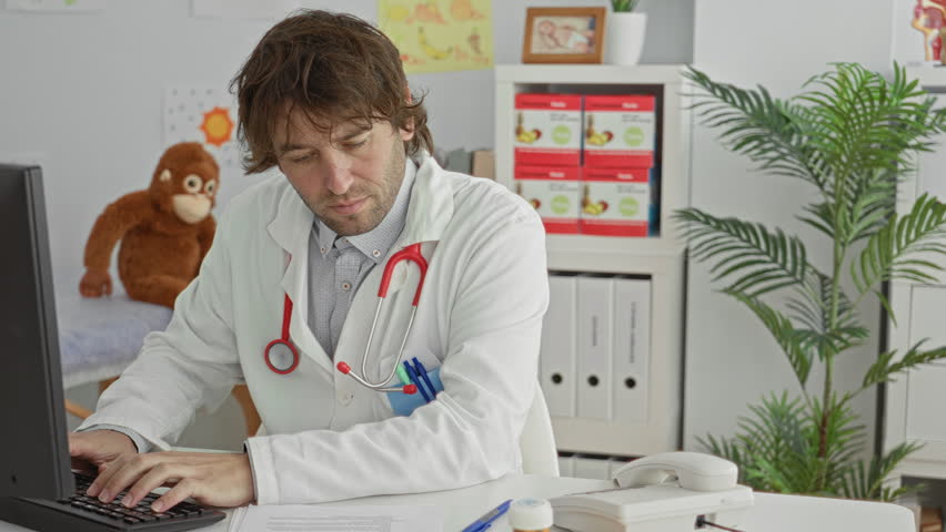 Man doctor types on keyboard with hands on keys at desktop computer, stethoscope draped on neck in clinic building; professional concentration.