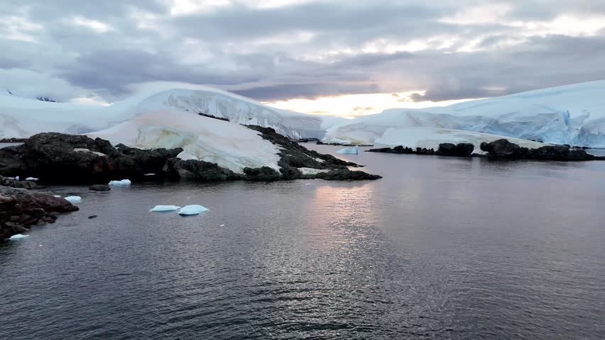 Cinematic aerial raising over the calm, dark waters of Foyn Harbour, revealing snow-covered volcanic islets and massive blue-layered ice shelves on the Antarctic Peninsula