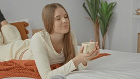 Young woman holding coffee mug with both hands, lying on bed in bedroom with knit throw, pillows, and potted houseplant by headboard; relaxation morning. - Powered by Shutterstock - Get 15% off with code: PIKWIZARD15