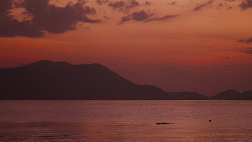 A bright deep red and orange beach sunrise overlooking the ocean with an island mountain in the background and purple clouds.