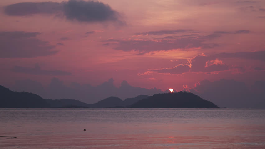 A sunrise over the calm ocean with the bright glowing sun peaking over the top of an island mountain with the orange and red skies reflecting in the water.