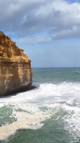 Aerial perspective of powerful white waves surging against rugged coastal cliffs under a cloudy sky.