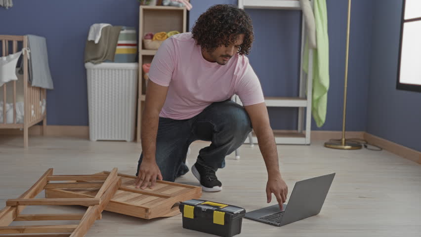 Man kneeling assembling cradle and reaching for parts in studio using laptop for instructions; frustration problem solving.