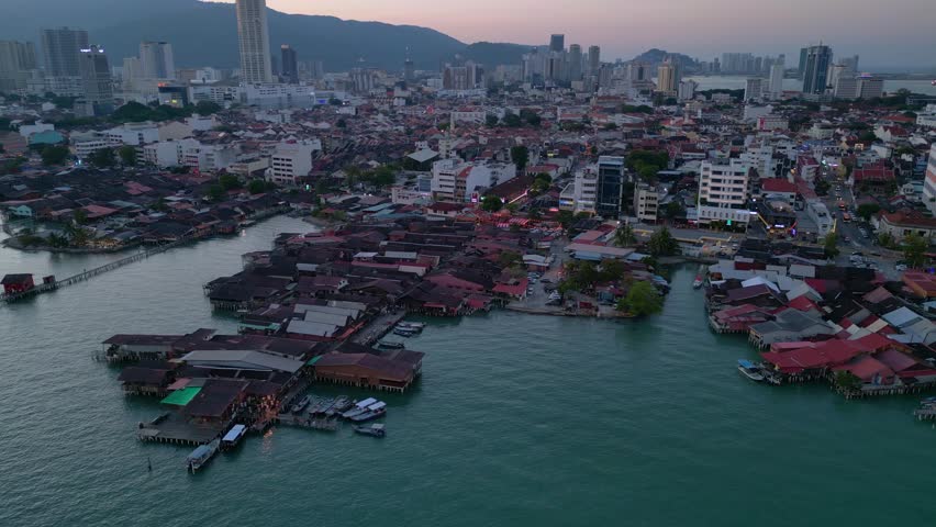Traditional stilt houses of George Town on the water, contrasting with the modern high rise buildings and urban skyline of Penang at sunset. Smooth aerial view wide orbit panorama overview drone
