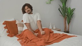 Man stretching arm while yawning on bed in bedroom with orange knit blanket, tassel pillows, rattan side table and hanging plants; relaxed. - Powered by Shutterstock - Get 15% off with code: PIKWIZARD15
