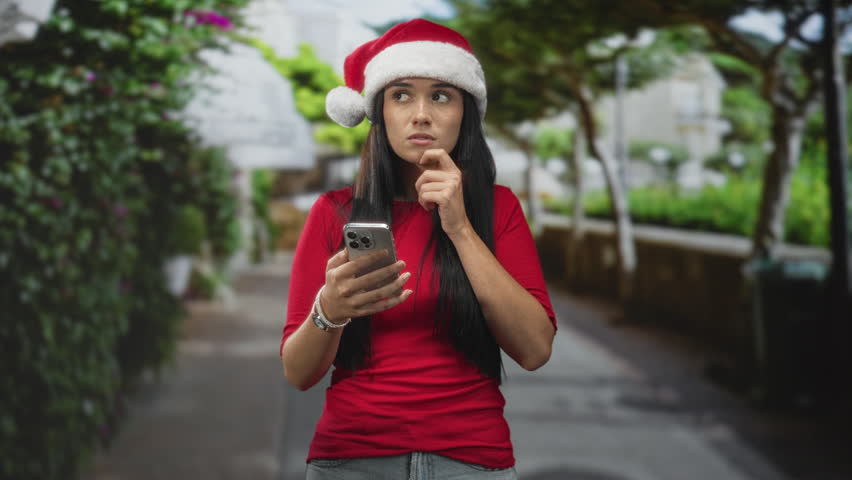 Young woman in red shirt and santa hat holding smartphone, hand to head adjusting hat on a street lined with trees; quiet contemplation.