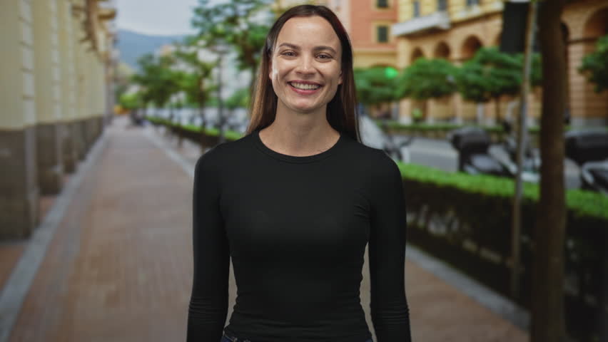 Woman points fingers and flashes thumbs up with open palms while smiling on a pedestrian city street lined with trees and buildings; playful approval.