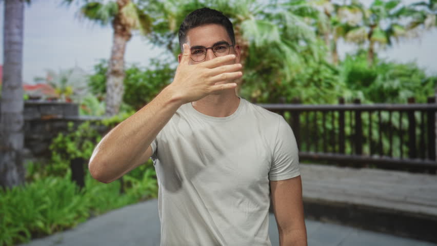 Man, young hispanic, wearing glasses and a white t shirt, placing hand on chest on a street outdoors near wooden railing; sincerity trust.