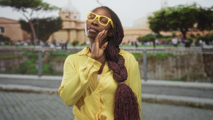 Woman with yellow glasses and long braids hand to chin at building plaza outdoor street scene; quiet contemplation.