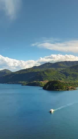 Cinematic aerial view of white boat sailing on deep blue lake water towards green mountain shoreline.