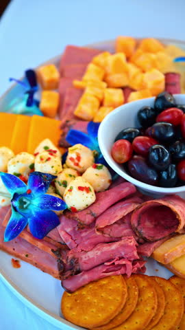 Charcuterie platter with cured meat cheese crackers and grapes. Gourmet appetizer tray featuring sliced ham, cheddar cheese, round crackers, and blue orchids on white plate.