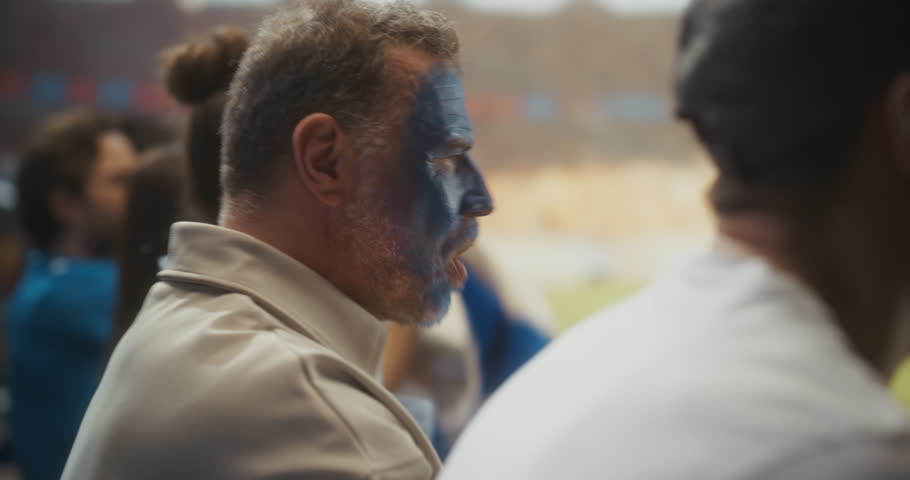 Close-up Portrait of a White Male Soccer Fan With Blue Painted Face Roaring in Celebration, Arms Raised Among Smiling Supporters at a Packed Stadium During a Pro League World Tournament Match.