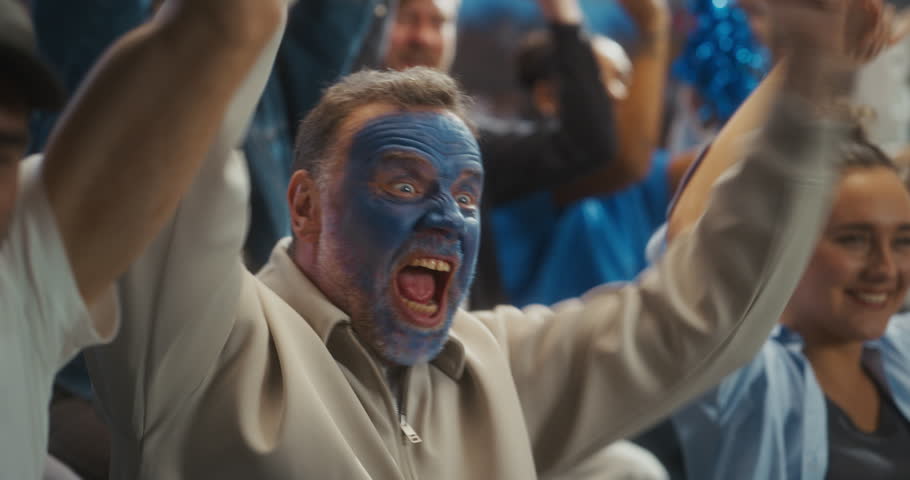 Close-up Portrait of a White Male Soccer Fan With Blue Painted Face Roaring in Celebration, Arms Raised Among Smiling Supporters at a Packed Stadium During a Pro League World Tournament Match.