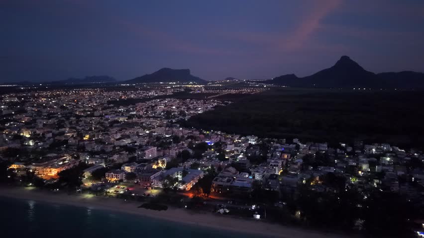 Coastal city at dusk, aerial perspective. Buildings glow softly along the shore. Beachfront stretches with gentle lights. Distant mountains frame the scene. No people visible