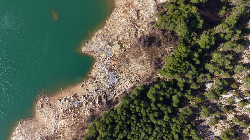 Top-down view of a receding lake shore revealing exposed rocky terrain and dry soil beside turquoise green water and dense forest