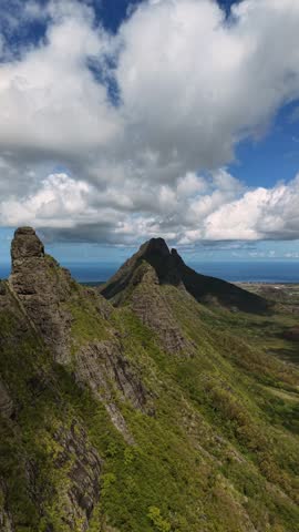 Mountain peak has green slopes and rocky terrain. Fluffy clouds drift over the bright blue sky. Distant ocean stretches to the horizon. Close-up shows mossy rock and ferns