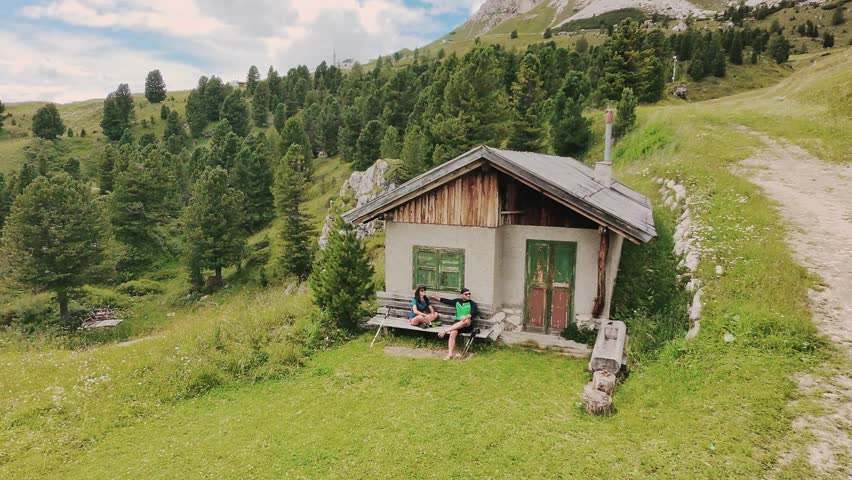 Aerial zoom in view couple fit hikers resting on bench outside a rustic stone hut in a lush alpine meadow in the Italian Dolomites. A scenic UNESCO World Heritage vista