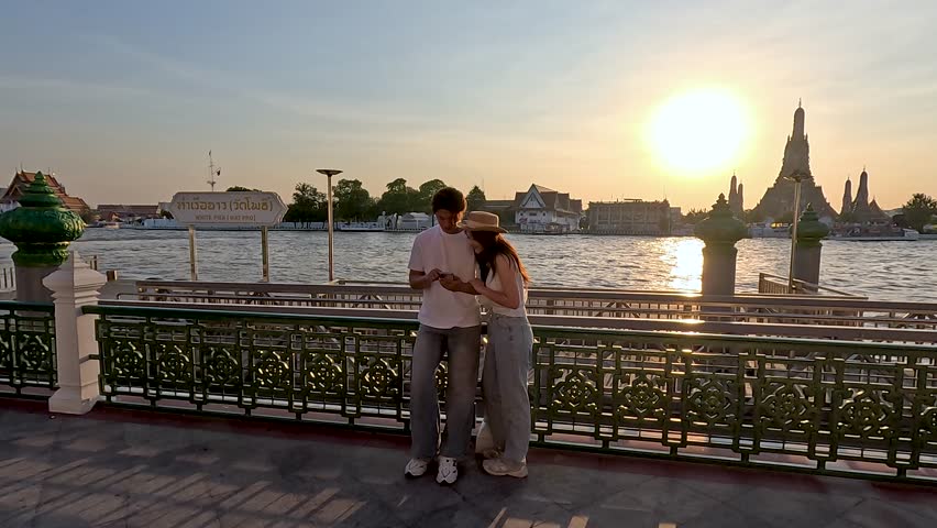 A couple stands on a riverbank walkway during golden hour with Wat Arun background