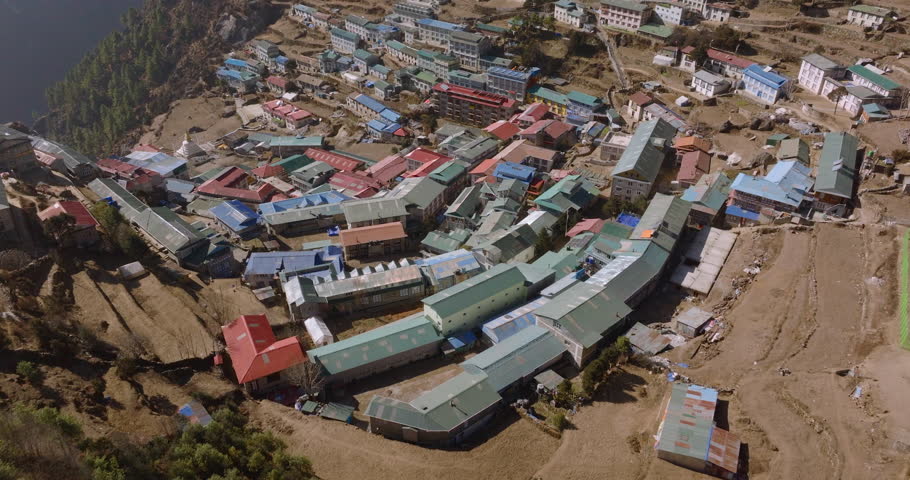 Aerial shot of Namche Bazaar showing birds flying over the amphitheater-shaped village with colorful rooftops, rugged hills and snow-capped Himalayan peaks under a clear blue sky in Nepal.