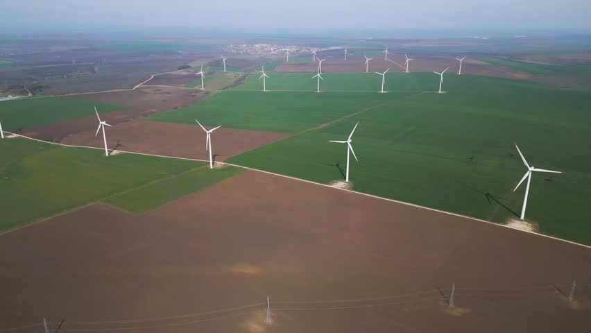High-angle drone shot of a large wind farm across green and brown fields. Features multiple turbines and high-voltage power lines, highlighting sustainable energy infrastructure and rural beauty.