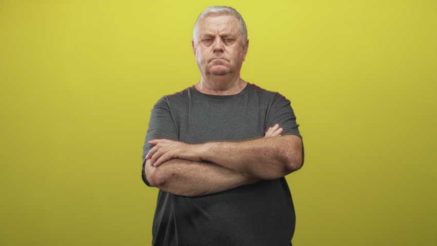 Man with hand on chin and folded arms in a studio against yellow backdrop, wearing a dark t shirt and frowning; quiet contemplation.