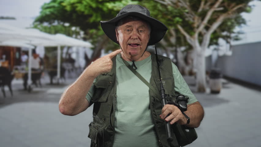 Senior man fisherman points finger to cheek, holding fishing rod, reel and binoculars while wearing hat and vest on street lined with trees and outdoor cafe; adventure curiosity.