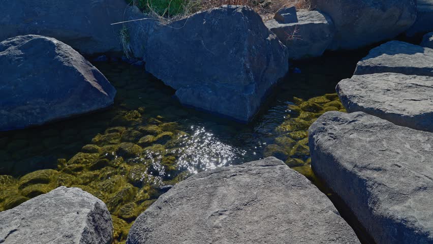 A serene, high-angle shot of clear water flowing between large, grey rocks, with bright sunlight creating shimmering reflections on the surface.