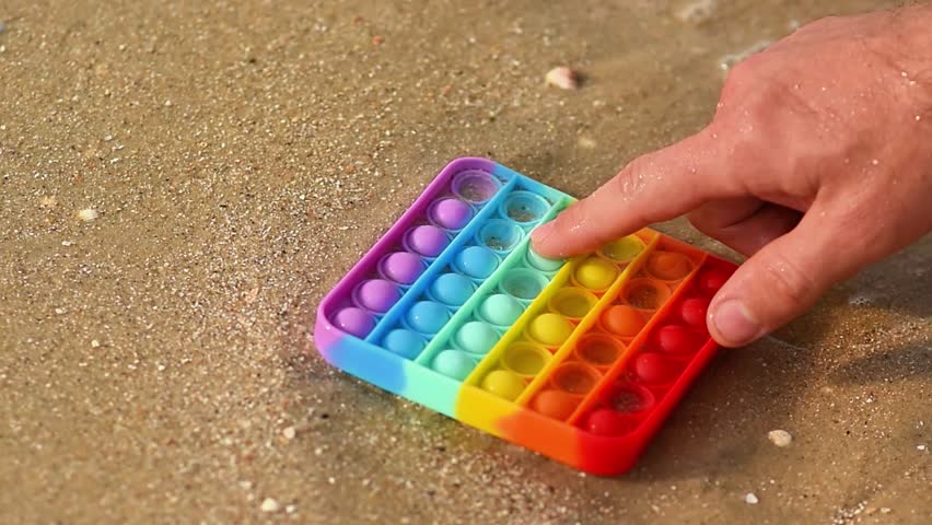 Person touching rainbow pop it on sand, bright silicone toy for sensory play, outdoor activity on beach, relaxation and fun content, ideal for kids or entertainment clips