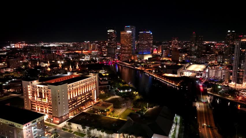 Aerial night view of downtown Tampa, Florida, with the Hillsborough River, illuminated skyline, bridges, and the iconic minarets of Plant Hall glowing beside the waterfront.