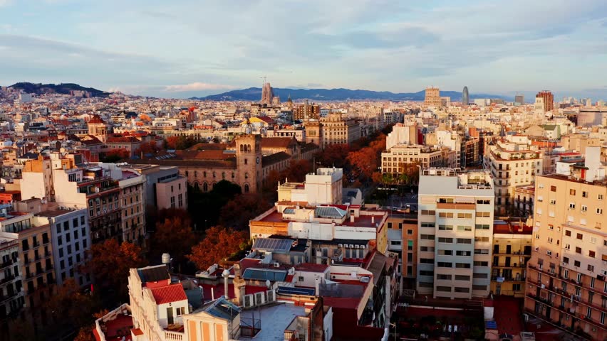 Aerial cityscape of Barcelona, Spain at golden hour, featuring dense rooftops, the Sagrada Familia and Torre Glories on the skyline with mountains in the background.