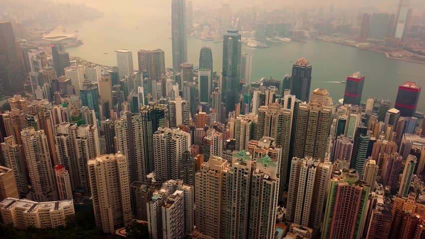 Aerial view of Hong Kong’s dense urban skyline, with Central and Wan Chai skyscrapers rising above Victoria Harbour in hazy daylight, showing modern architecture and waterfront city life.