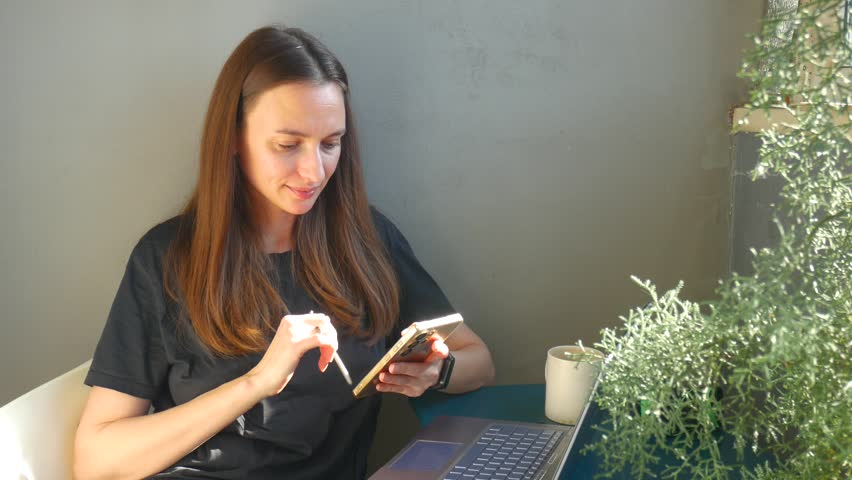 Young woman using smartphone and stylus pen while sitting at desk with laptop. Caucasian lady working online or browsing the internet in a bright modern office.