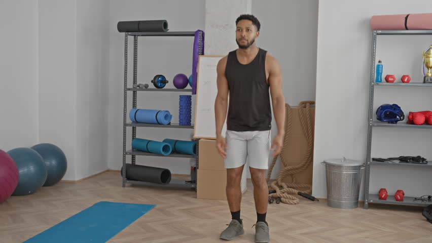 Man in black tank top and white shorts raises arms overhead performing a jumping jack on a blue mat in a fitness studio with exercise balls and ropes visible; energy fitness focus.