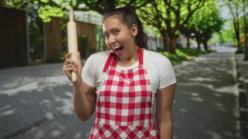 Woman holding rolling pin on a tree lined street wearing red checkered apron and smiling broadly; home baking joy.