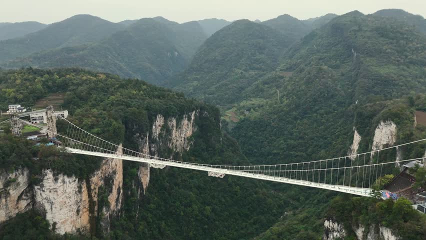 Crossing glass suspension bridge in Zhangjiajie, China. Transparent walkway crosses lush mountain canyon. Suspended glass bridge connecting cliffs over misty valley