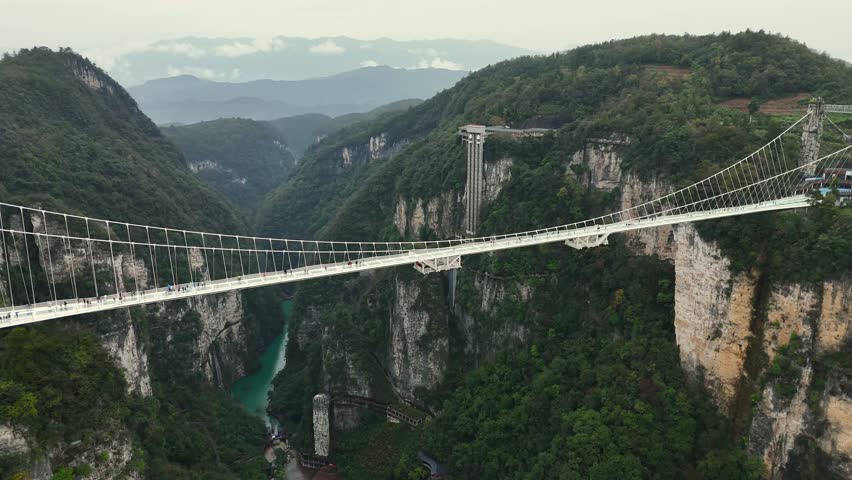 Panoramic shot showcasing towering bridge spanning deep forested canyon with scenic river below. Crossing glass suspension bridge in Zhangjiajie, China