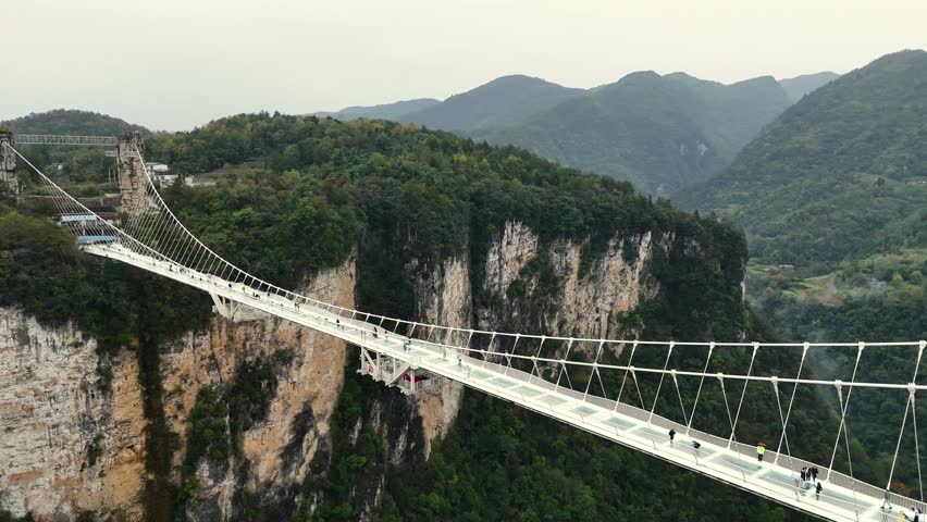 Adventurers carefully cross glass pathway spanning mountain valley with scenic and uncertain surroundings. Crossing glass suspension bridge in Zhangjiajie, China