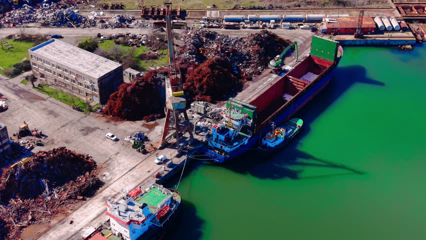 Industrial crane loading recycled metal into the hold of a large blue and red bulk carrier at a harbor pier. Aerial view of scrap metal loading onto cargo ship at port.