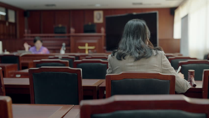 Woman sits in courtroom, facing judge. Back view shows her attentive posture. Room has wood paneling, rows of chairs. Judge sits at raised bench, behind large screen
