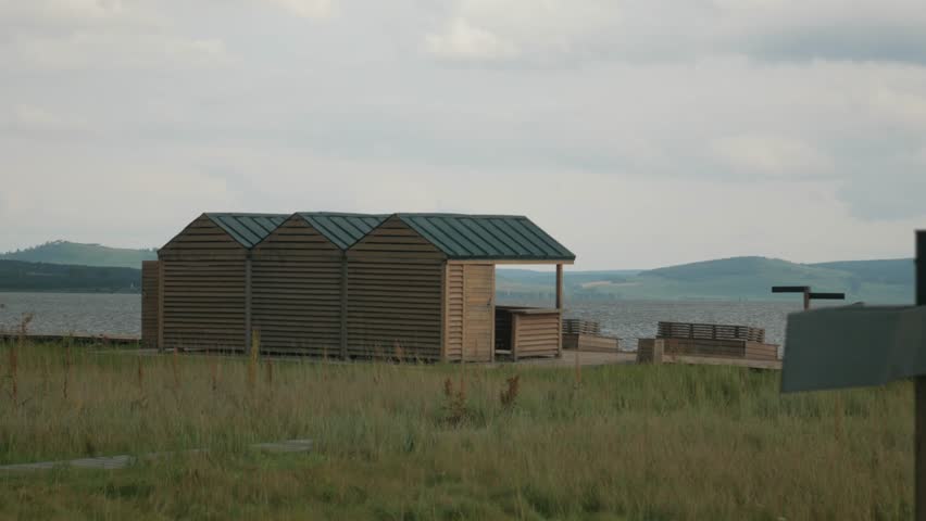 Wooden Cabins Static Lakeshore Handheld Shot. A static handheld shot of small wooden cabins standing on a grassy clearing near a lake.